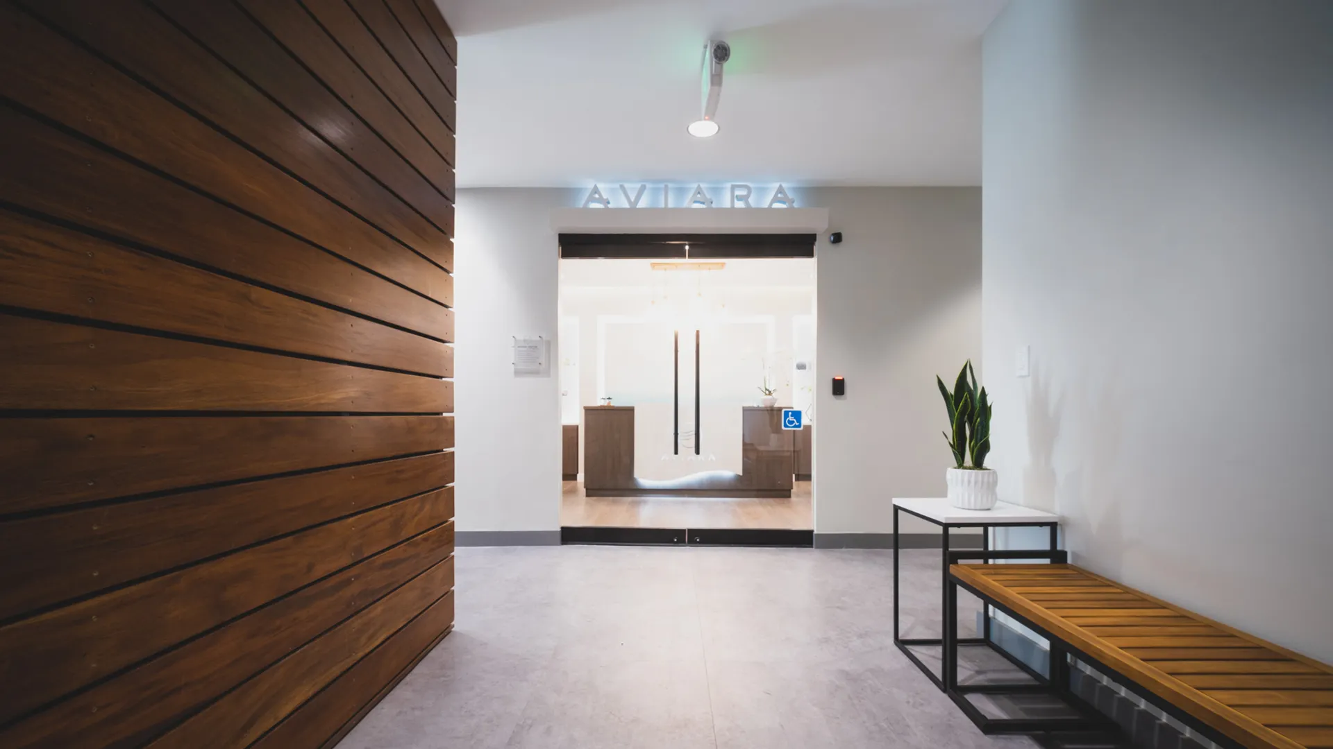 Entrance hallway at Aviara Centers in Los Alamitos with wood accent wall, bench seating, and glass doors beneath illuminated Aviara signage